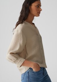 Beige button-up shirt with a relaxed fit, textured surface, V-neck, and 3/4 sleeves, paired with blue jeans.