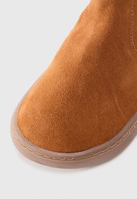 Botte de cheville en suede marron avec un bout arrondi et une semelle en caoutchouc beige avec des détails de couture le long du bord. Texture lisse sur l'ensemble.