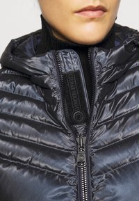 Tummansininen toppatakki kiiltävällä pinnalla, jossa on vetoketjullinen etuosa, korkea kaulus ja brändätty merkki vetoketjulinjan varrella.