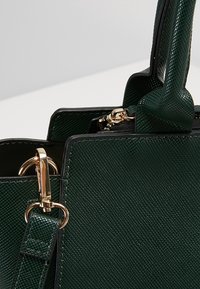 Sac à main en cuir texturé vert foncé avec des ferrures métalliques dorées et des poignées structurées sur un fond neutre.