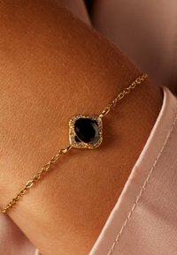Gouden armband met een vierkante zwarte steen omringd door kleine heldere stenen, bevestigd met een delicaate ketting op een gebruinde huid achtergrond.