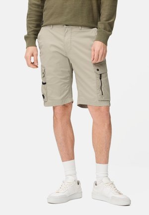 Mann trägt beigefarbene Cargoshorts, weiße Socken und weiße Turnschuhe, die Hände entspannt an den Seiten vor einem einfarbigen Hintergrund.