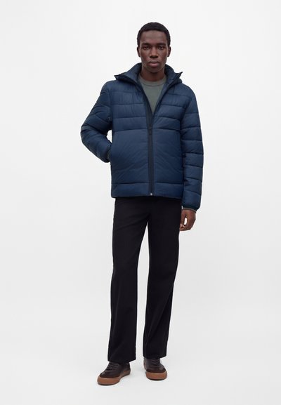 Homme debout portant une doudoune bleu marine, un pantalon foncé, des chaussures marron et une chemise verte sur un fond blanc uni.