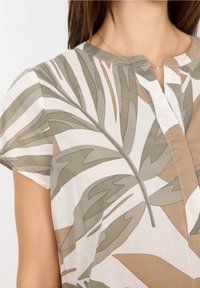 Blusa leggera a maniche corte in bianco sporco con motivi di foglie verdi e beige, caratterizzata da un collo senza bottoni e una vestibilità morbida.