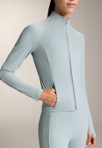 Svijetloplava, uska jakna s dugim rukavima i rebrastom teksturom, visokim ovratnikom i prednjim zatvaračem; ima bočne džepove i bezšavnu konstrukciju.