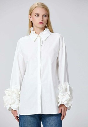Chemise blanche à boutons avec un col haut et des manches longues, ornée d'accents volants surdimensionnés aux poignets et d'une texture en coton douce.