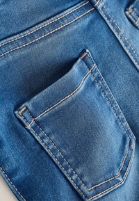 Jeans en denim bleu moyen avec une texture délavée. Présente une poche plaquée avec des coutures contrastées en tan et un design propre et classique.