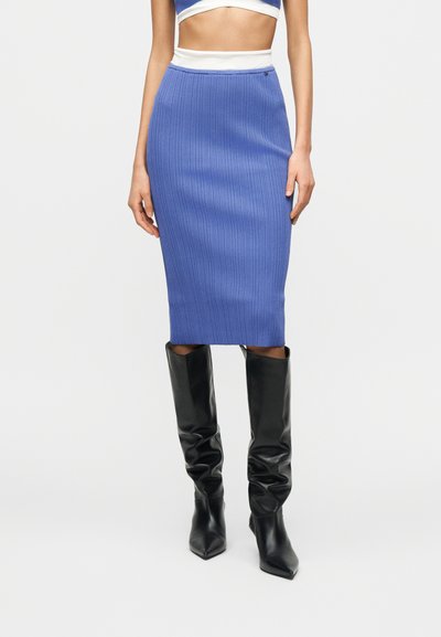 Femme portant une jupe midi bleue côtelée associée à des bottes noires à bouts pointus montant jusqu'aux genoux, debout devant un fond uni.