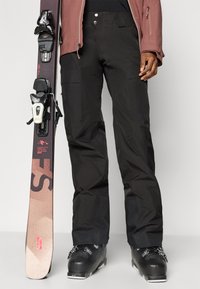 Μαύρες σκιέρ παντελόνες με υφή, συνδυασμένες με μαύρες σκιέρ μπότες. Ένα σκι με ροζ και καφέ σχέδιο κρατιέται δίπλα.