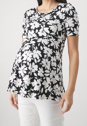 Zwangere persoon die een zwart-wit gebloemd zwangerschapshemd met korte mouwen en een witte broek draagt, poserend tegen een effen achtergrond.