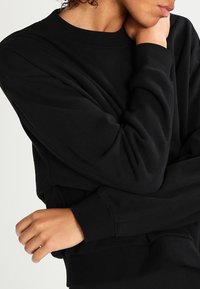 Sweat-shirt noir avec un col rond et des manches longues, présentant une coupe décontractée et un tissu doux et texturé. Détail de couture subtil au niveau des poignets.