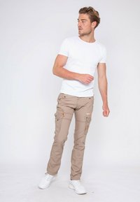 Pantalons cargo beiges avec plusieurs poches et des accents orange, associés à un t-shirt blanc uni et des baskets blanches. Texture de tissu lisse.