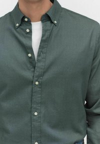 Man in een donkergroen overhemd met knopen over een wit shirt met ronde hals, met de bovenste knoop los.