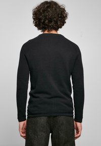 Svart långärmad tröja med en texturerad stickad design. Ribbad mudd och nederkant. Bärs med mörkgrå byxor; visas bakifrån.