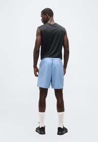 Άνδρας που στέκεται με την πλάτη προς τα εμπρός, φοράει μαύρο αμάνικο πουκάμισο, ανοιχτό μπλε σορτς, λευκές κάλτσες και μαύρα αθλητικά παπούτσια, σε ένα απλό φόντο.