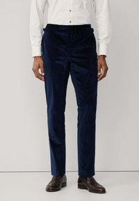 Muškarac u tamnoplave barske hlače, bijelu košulju dugih rukava i tamnosmeđe kožne mokasine, stoji pred jednostavnom pozadinom.