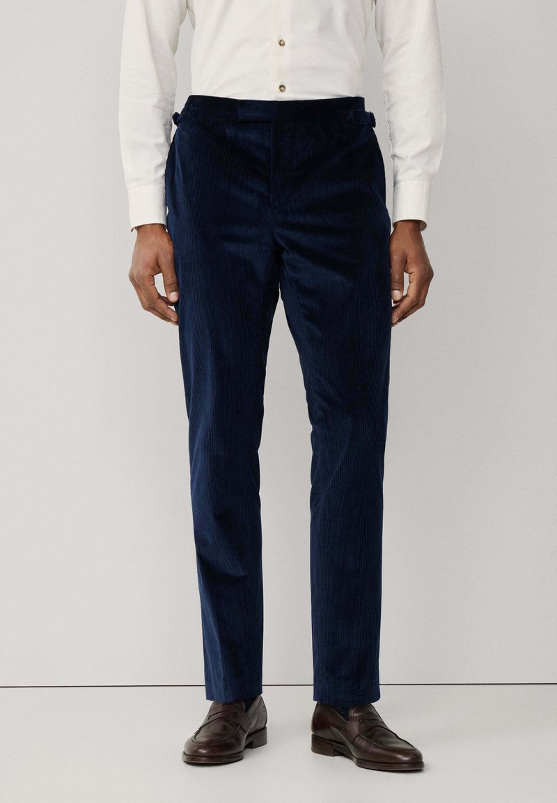 Muškarac u tamnoplave barske hlače, bijelu košulju dugih rukava i tamnosmeđe kožne mokasine, stoji pred jednostavnom pozadinom.