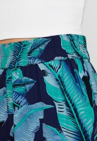 Pantalon à imprimé floral sur fond bleu marine, avec de grandes feuilles tropicales vertes. Taille élastique avec tissu texturé.