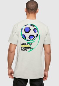 Svetlo béžové tričko s veľkým, farebným grafickým motívom futbalovej lopty s čiernymi, modrými a zelenými akcentmi. Text znie "Športový nedeľný futbalový klub."