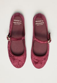 Un par de zapatos Mary Jane de terciopelo burdeos con correas de hebilla doradas y "Woman by Fred Martin Made in Spain" impreso en las plantillas.