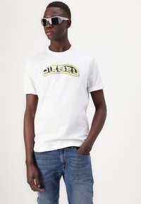White cotton t-shirt with a yellow and black "DIESEL" logo, paired with blue denim jeans and gray sunglasses.