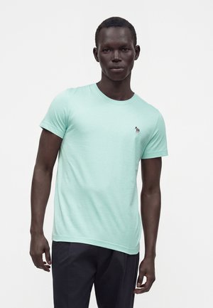 Mann, der ein hellgrünes kurzärmeliges T-Shirt mit kleinem Zebra-Logo auf der Brust und dunkle Hose vor einem schlichten weißen Hintergrund trägt.