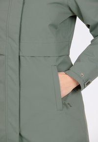 Ljusgrön vattentät jacka med rak skärning, framdragkedja, sidofickor och tryckknappar på ärmarna. Slät, matt textur.