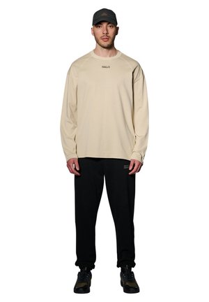 Beige langwerpig shirt met textuurstof en "HALO" logo, gecombineerd met zwarte trainingsbroek met een kleine graphic. Zwarte pet en schoenen.