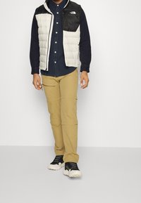 En pufferväst i vitt och svart, långärmad marinblå skjorta, beige cargo-byxor och svarta sneakers med vita sulor. Avslappnad utomhusklädsel.