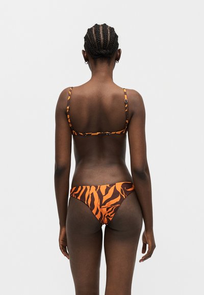 Γυναίκα με πλεγμένα μαλλιά στέκεται με την πλάτη, φοράει πορτοκαλί και μαύρο μαγιό με ρίγες τίγρης σε απλό λευκό φόντο.