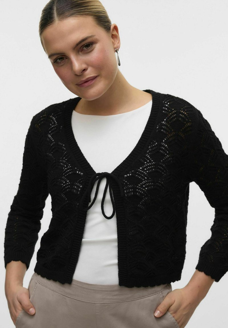 Cardigan corto nero con un motivo simile a pizzo, scollo a V e lacci sul davanti. Texture morbida con maniche lunghe e bordi a costine.