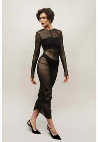 Czarna, prześwitująca sukienka z długim rękawem, z marszczeniami i dopasowanym krojem, z wysokim dekoltem i długością do połowy łydki, zestawiona z czarnymi obcasami.