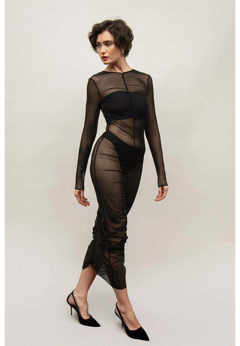 Czarna, prześwitująca sukienka z długim rękawem, z marszczeniami i dopasowanym krojem, z wysokim dekoltem i długością do połowy łydki, zestawiona z czarnymi obcasami.