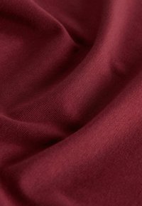 Bordowy materiał o miękkiej teksturze, ukazujący drobne prążkowanie i delikatny połysk, miękko układający się w fałdy, podkreślający swoją elastyczność.