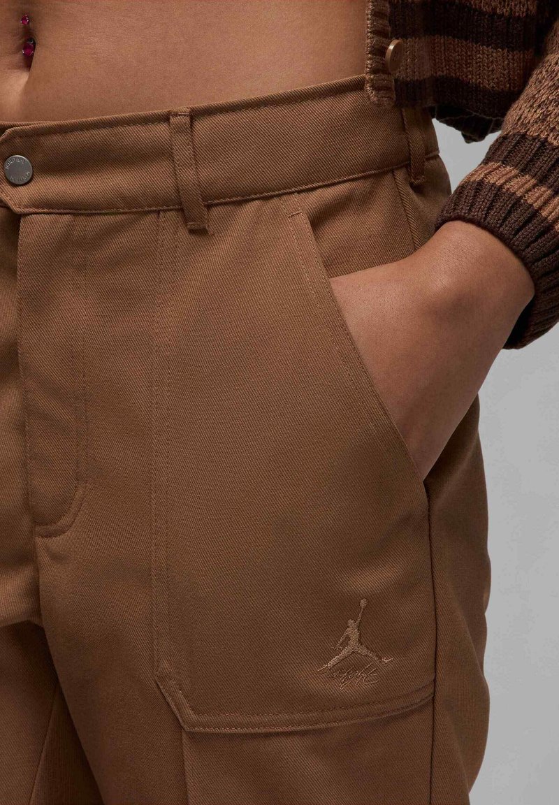 Pantaloni cargo marroni realizzati in un tessuto robusto con una superficie testurizzata, dotati di una tasca laterale e di un logo in rilievo discreto sulla gamba inferiore.
