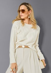 Frau trägt einen cremefarbenen Strickpullover, beigefarbene, plissierte Hose, einen goldenen Gürtel, große Ohrringe, übergroße Sonnenbrille und hält eine cremefarbene Clutch-Tasche.