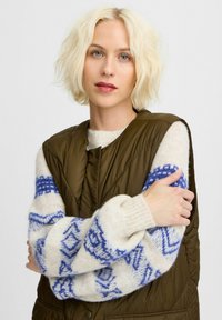 Olivengrüne gepolsterte Weste über einem cremefarbenen Pullover mit blauen geometrischen Mustern; weiche Textur und das gesteppte Design sind eindeutig sichtbar.