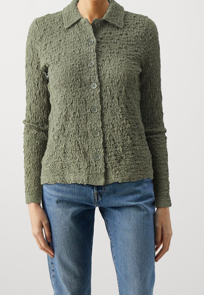 Femme portant une chemise texturée vert olive à boutons et manches longues ainsi qu'un jean en denim bleu sur un fond uni.