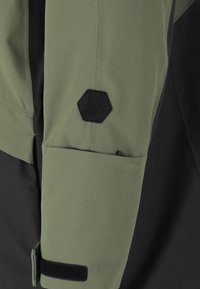 Der Ärmel der grün-schwarzen Jacke verfügt über einen strukturierten, hexagonalen Patch. Der Stoff ist glatt und strapazierfähig, mit einer sicheren Tasche und einem Klettverschluss.