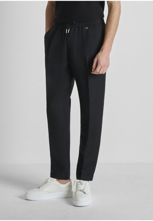 Hombre con pantalones negros con cordón y bolsillos laterales, y zapatillas blancas bajas, de pie frente a un fondo gris liso.