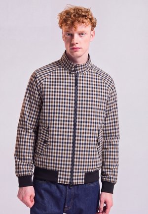 Junger Mann mit lockigem roten Haar, der eine beige-braun-schwarz karierte Jacke mit Reißverschluss und dunkelblaue Jeans vor einem einfarbigen Hintergrund trägt.