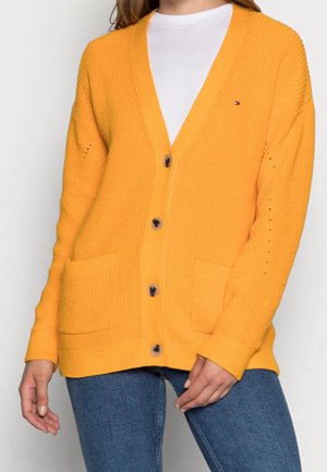 Cardigan tricoté jaune avec un décolleté en V profond, de grands boutons, des poches avant et une texture côtelée. Porté sur une chemise blanche et un jean bleu.
