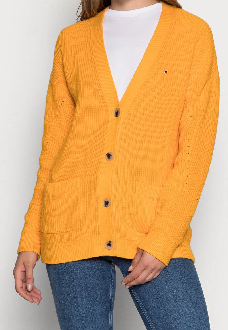 Keltainen neulottu neuletakki, jossa on syvä V-kaula-aukko, isot nappit, etutaskut ja ribbitetty rakenne. Päällä valkoinen paita ja siniset farkut.