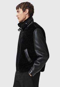 Black shearling vest with leather sleeves, high collar, and snap closure. Features a side pocket and ribbed cuffs. Smooth leather texture.