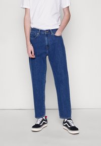 Blue denim jeans with a straight-leg fit, five-pocket design, and stitched accents, paired with black and white sneakers.