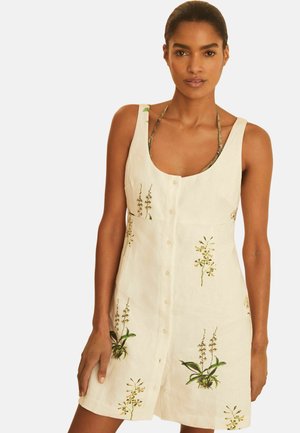 Kvinde iført ærmeløs beige kjole med knap foran og grøn botanisk print, stående mod en ensfarvet baggrund.