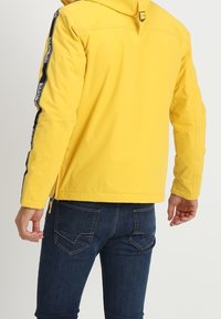 Giacca gialla con cappuccio, dotata di strisce nere sui lati, realizzata in materiale liscio e resistente all'acqua. Abbinata a jeans scuri.