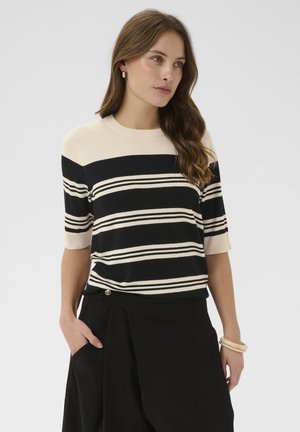 Kobieta o długich brązowych włosach, ubrana w czarny i beżowy sweter w paski z krótkim rękawem oraz czarną spódnicę, z ręką w kieszeni i bransoletkami na nadgarstku.