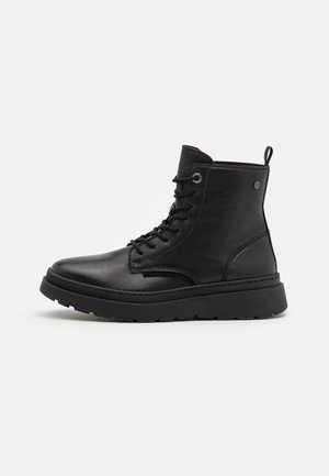 Bottine en cuir noir avec semelle épaisse, devant lacé, languette au talon et œillets métalliques, présentée en profil sur fond blanc.