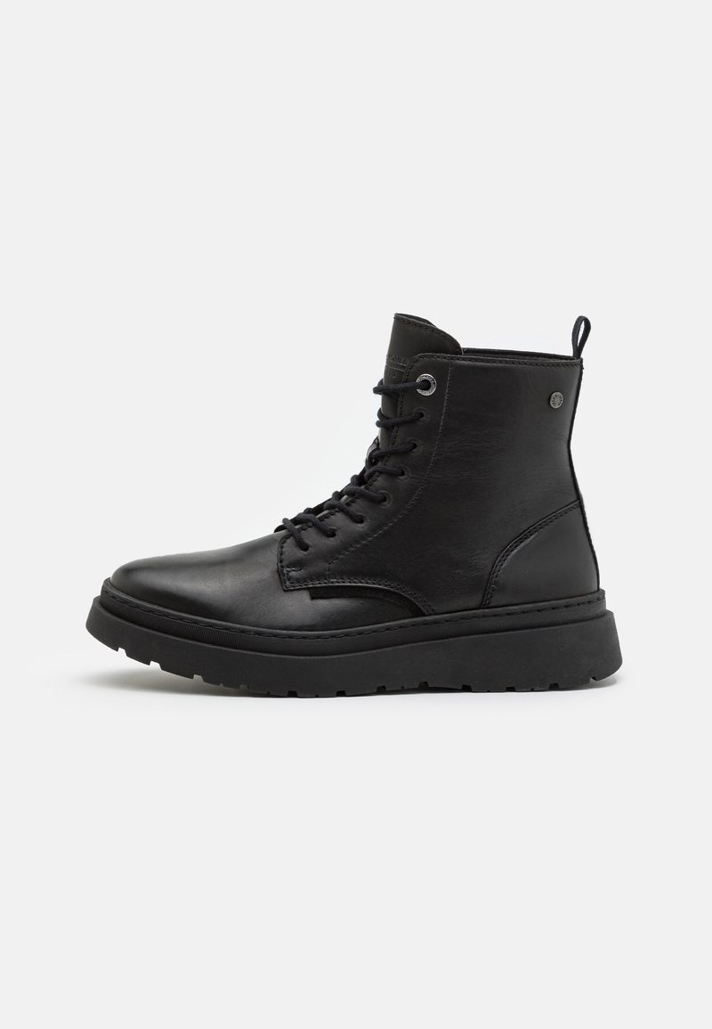 Bottine en cuir noir avec semelle épaisse, devant lacé, languette au talon et œillets métalliques, présentée en profil sur fond blanc.